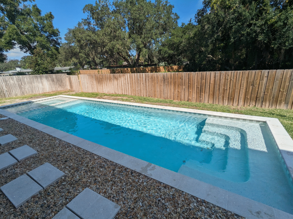 Minimalist rectangle pool with light mosaic tile and oversized coping, set against a wood privacy fence 