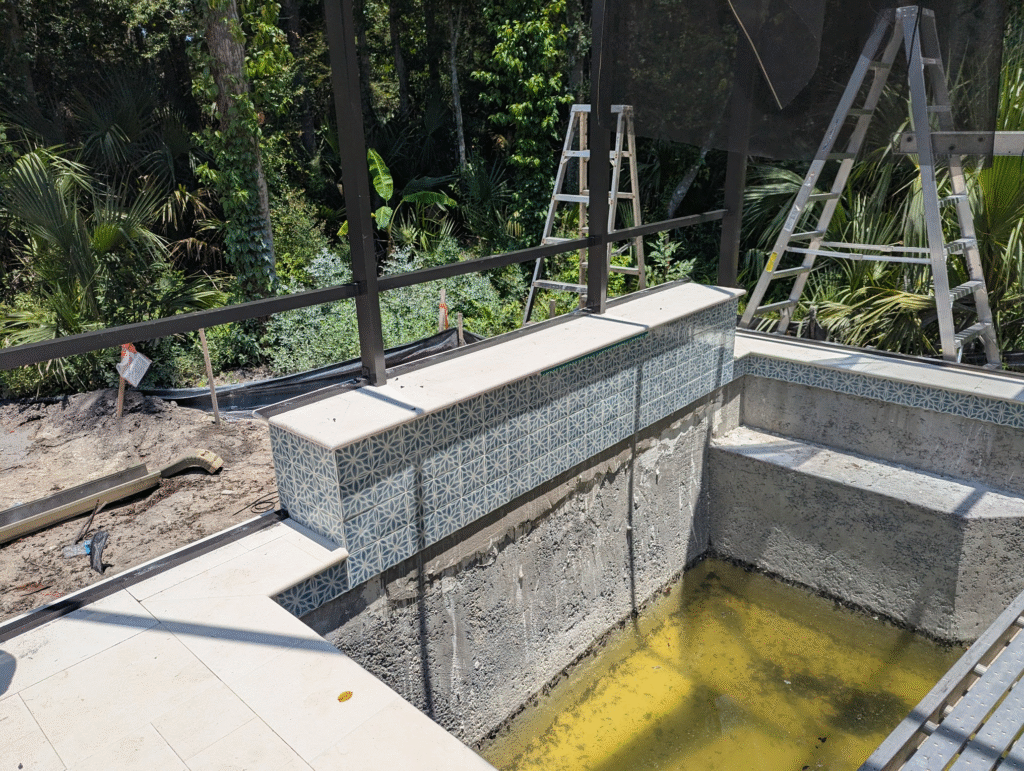 Installation of patterned mosaic tile on a raised wall beam during pool construction, showcasing a bold geometric design