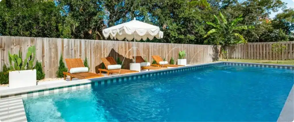 View of contemporary pool with teak loungers, umbrella shade, deck jets, and warm wood privacy fence